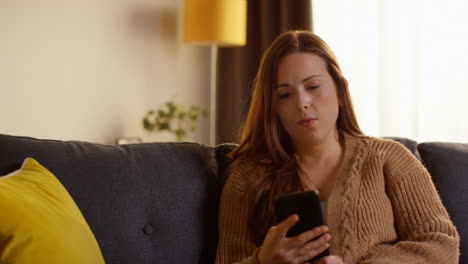 Puzzled-Woman-Sitting-On-Sofa-At-Home-Answering-Cold-Or-Nuisance-Call-On-Mobile-Phone-That-She-Doesn't-Recognise